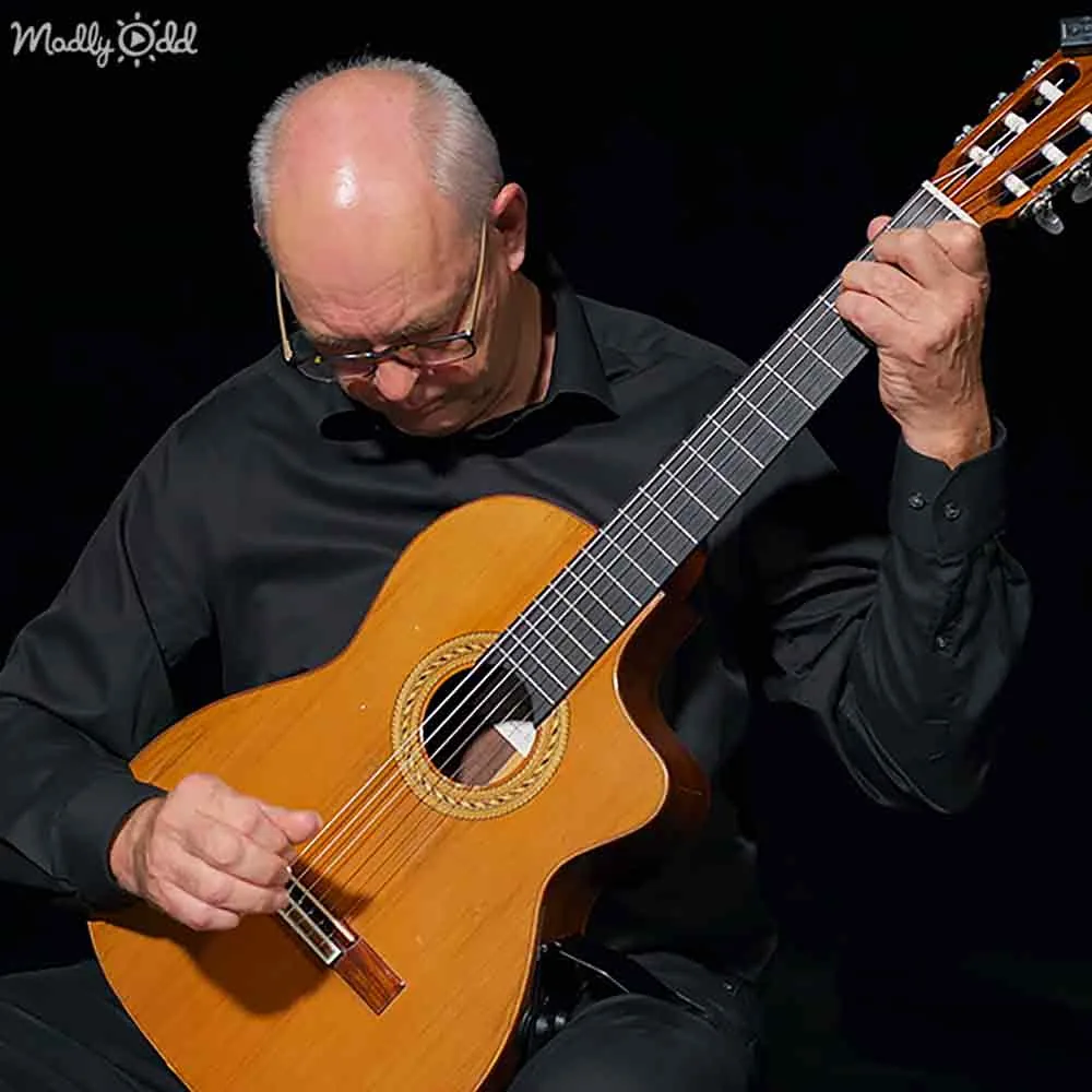 A man in a black shirt plays a classical guitar with a focused expression.