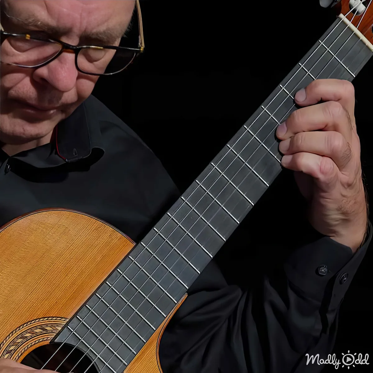 A man in glasses plays a classical guitar, focusing intently on the strings.