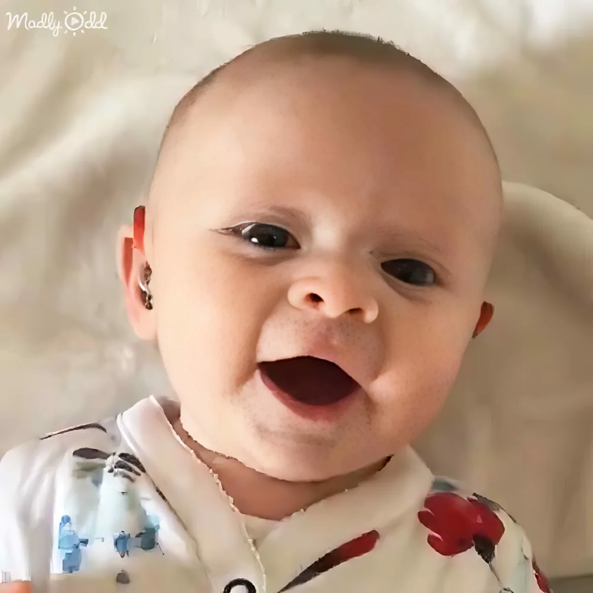 A baby with a bald head, wearing a white outfit with red and blue patterns, smiles widely.
