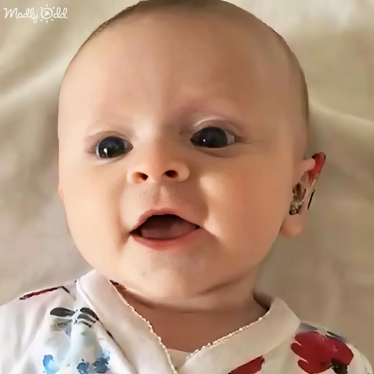 A baby with a shaved head, wearing a white and red outfit, is smiling and looking directly at the camera.