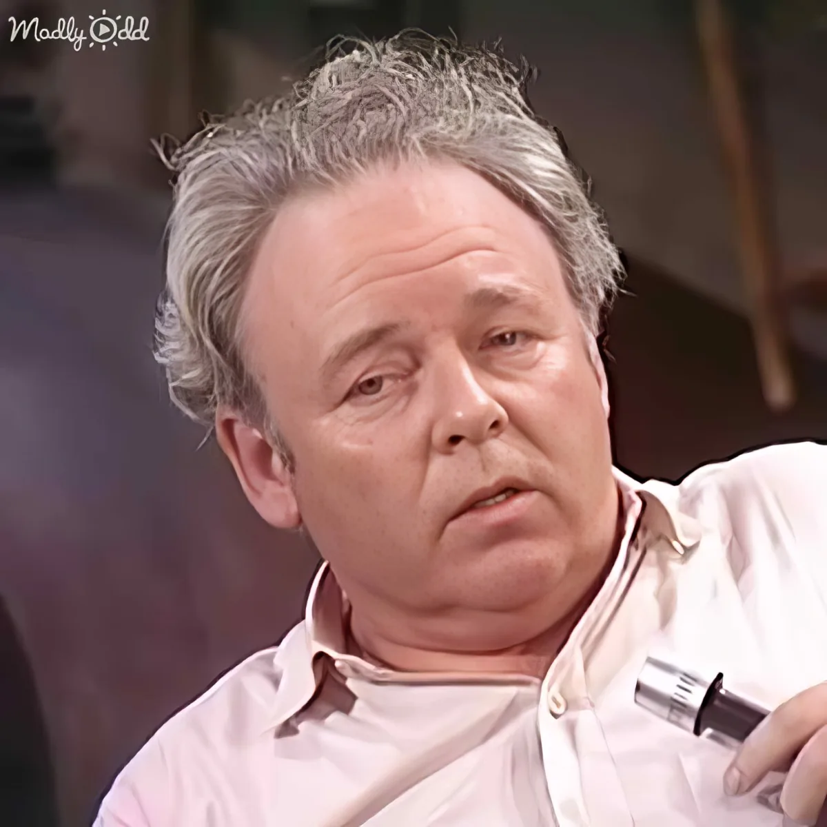 A man with gray hair, wearing a light pink shirt, holding a microphone, appears to be speaking or reacting to something.