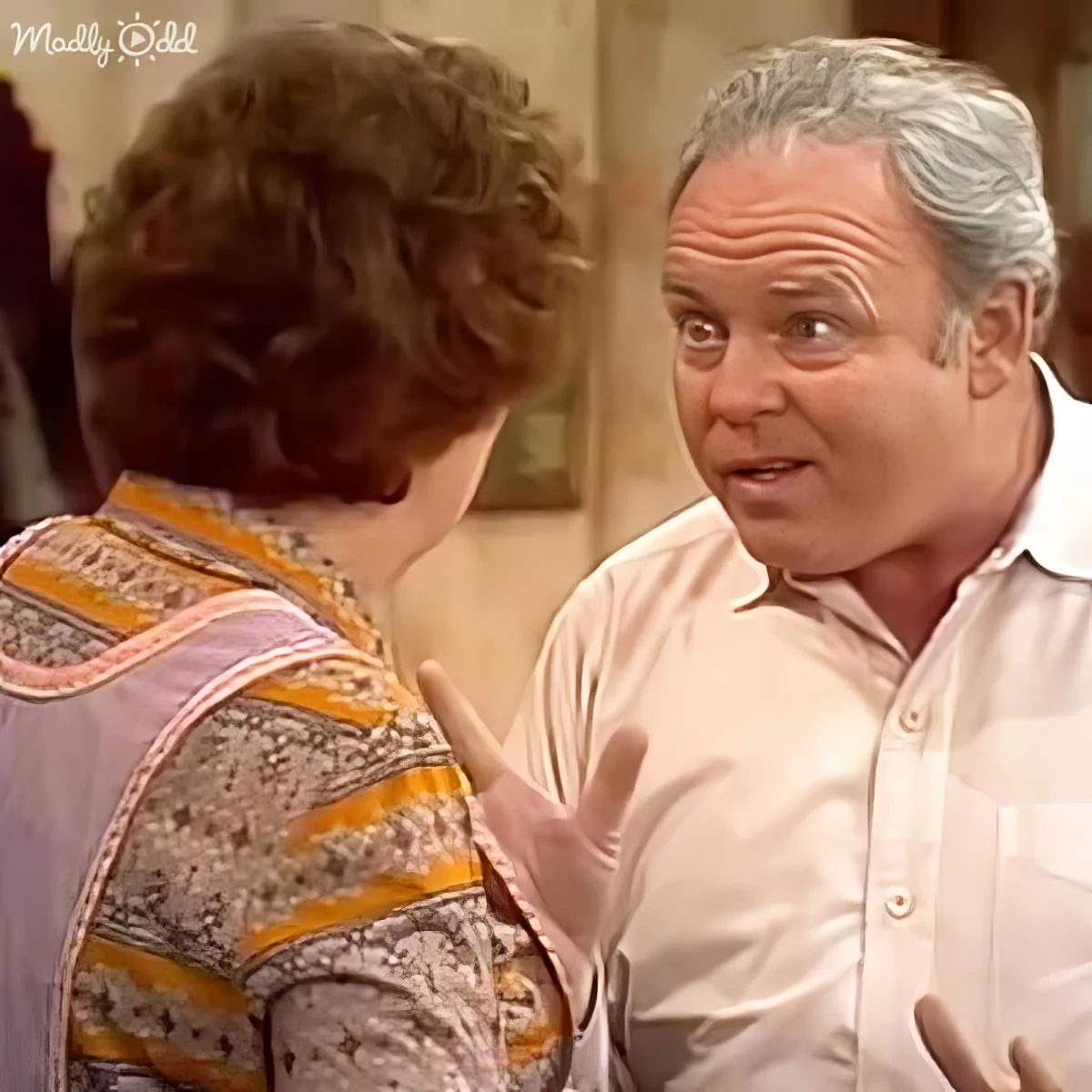 A man in a white shirt gestures while talking to a woman in a patterned dress in a room.