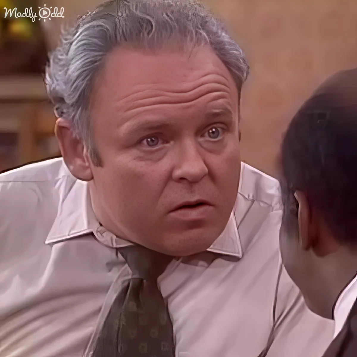 A man in a white shirt and tie leans forward, looking surprised or concerned, while another person is partially visible...
