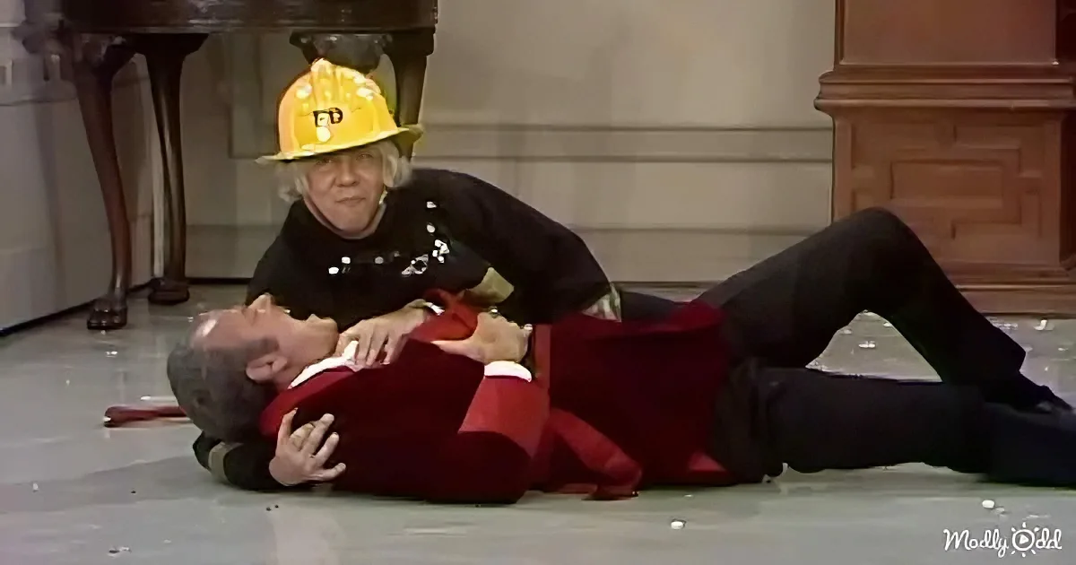 A man in a red suit is lying on the floor, being held down by another person wearing a firefighter helmet.