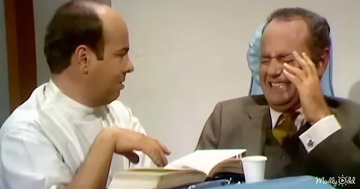 A man in a white shirt is sitting at a table, looking at another man in a suit who is laughing and covering his face.