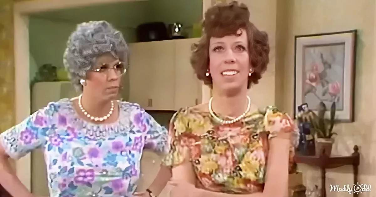 Two women in floral dresses, pearls, and 1970s hairstyles stand in a kitchen, one with hands on hips.