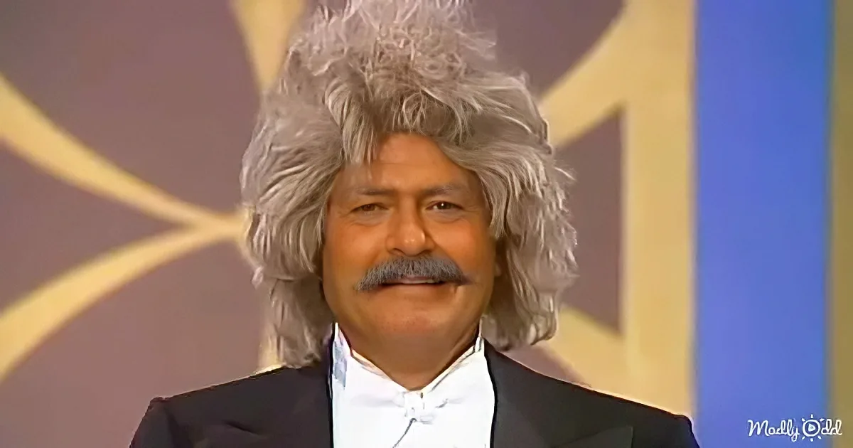 A man with long, gray, curly hair and a mustache, wearing a black suit and white shirt, stands in front of a blurred...