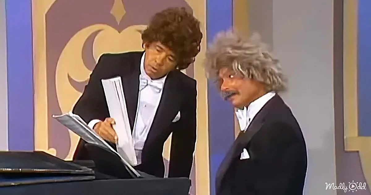 A man in a tuxedo holding papers, another man in a suit with a mustache beside him.