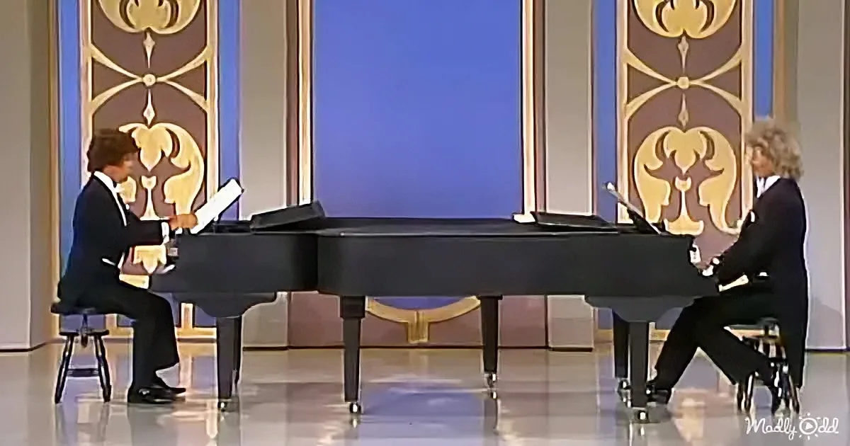 Two pianists in suits play a grand piano on a stage with ornate gold and blue decorations.