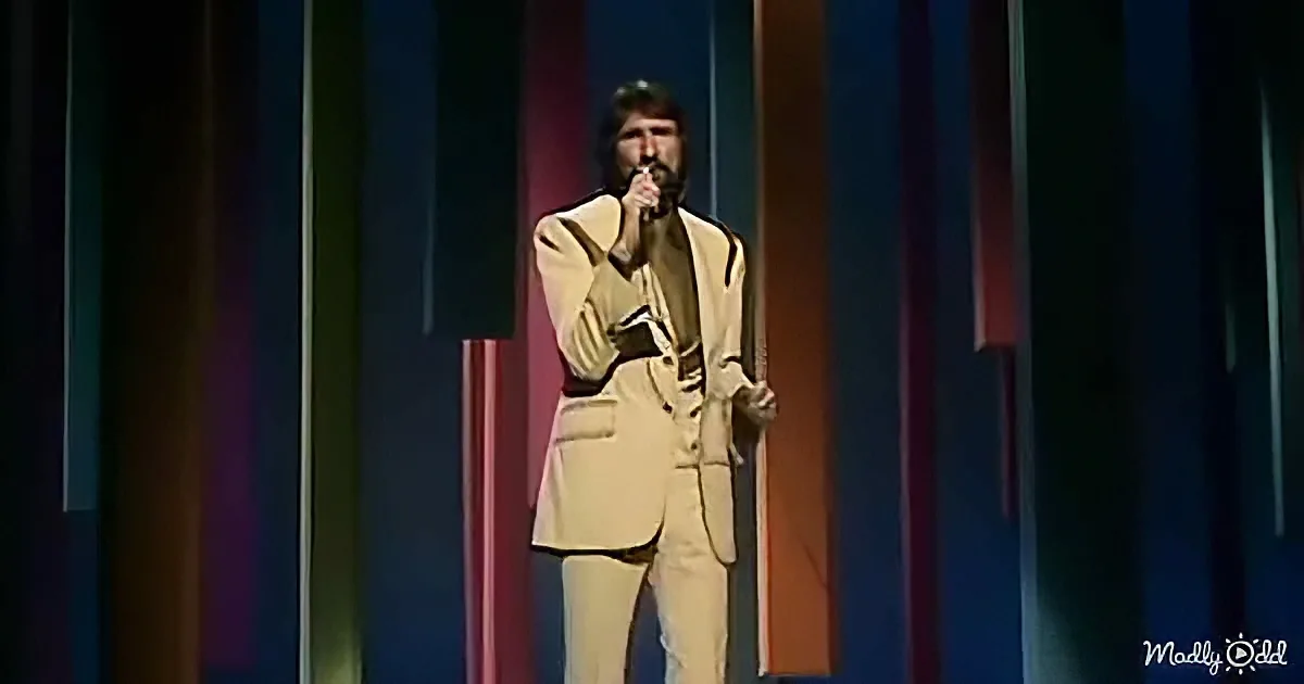 A man in a beige suit sings into a microphone on stage with vertical colored stripes.