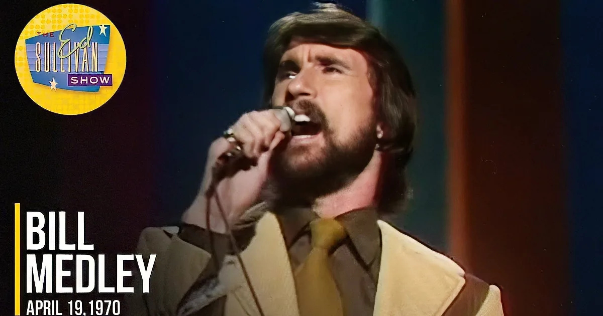 A man with long hair and a beard sings into a microphone, wearing a yellow jacket and tie.