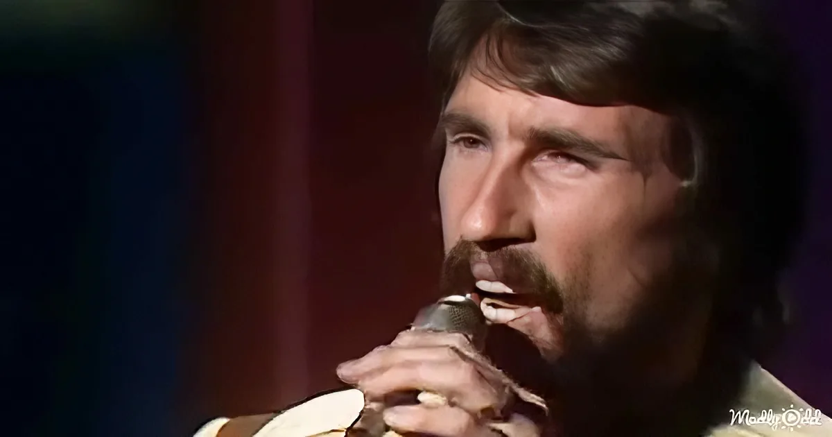 A man with a mustache and beard sings into a microphone, wearing a light-colored shirt.
