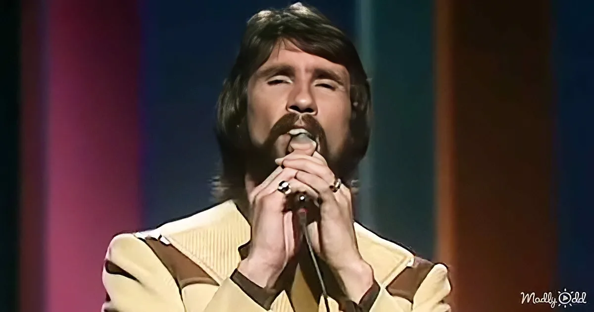 A man with a mustache and long hair sings into a microphone, wearing a yellow and brown striped shirt.