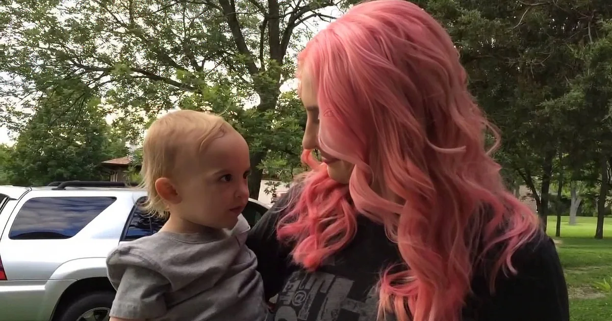 A child with blonde hair interacts with a person with pink hair in an outdoor setting.