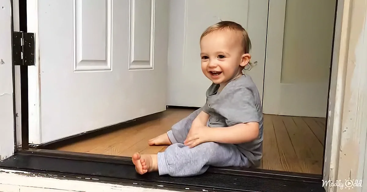 A smiling baby sits on a door threshold, wearing a gray outfit, with a white door and wooden floor in the background.