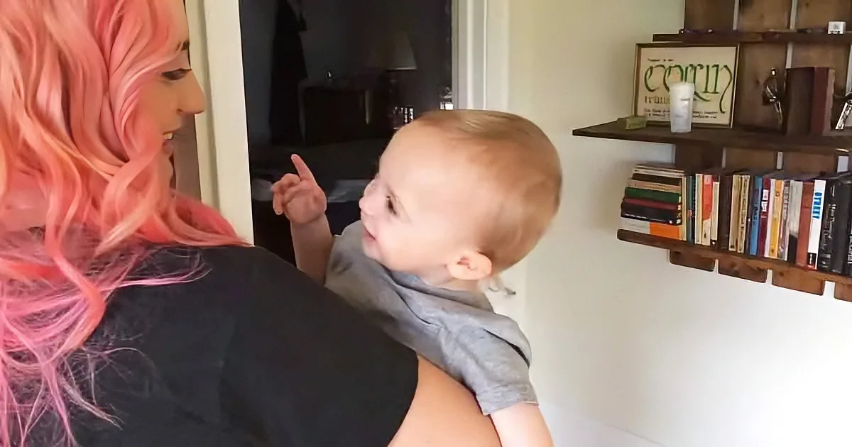 A woman with pink hair holds a baby, both looking at each other in a cozy room with shelves.