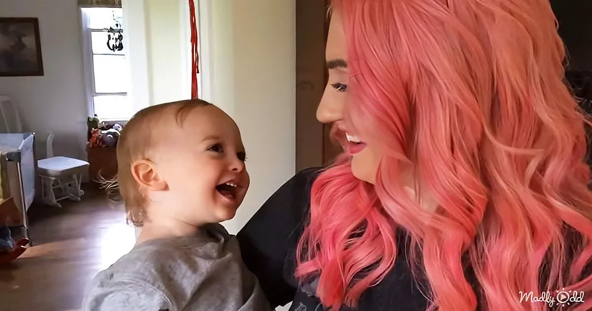 A woman with vibrant pink hair smiles at a laughing baby in a cozy room.