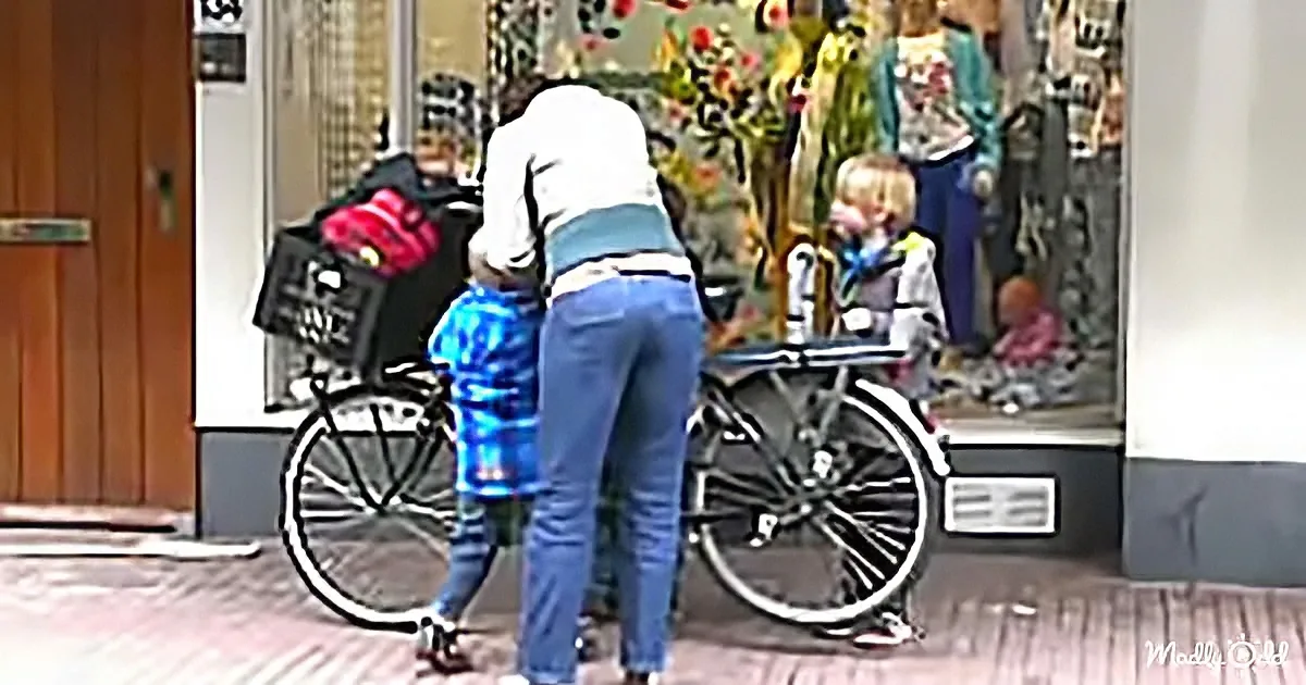 A woman helps a child onto a bicycle in front of a store window.