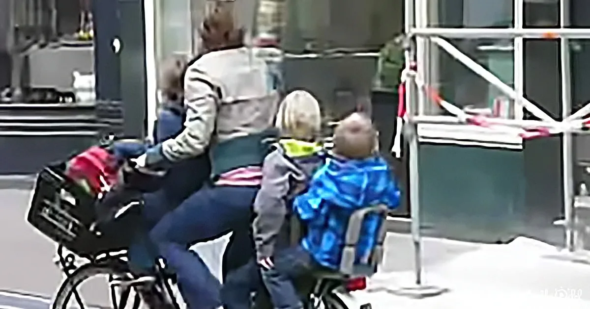 A woman rides a bicycle with two children in a city setting, one in a front basket and one in a rear child seat.