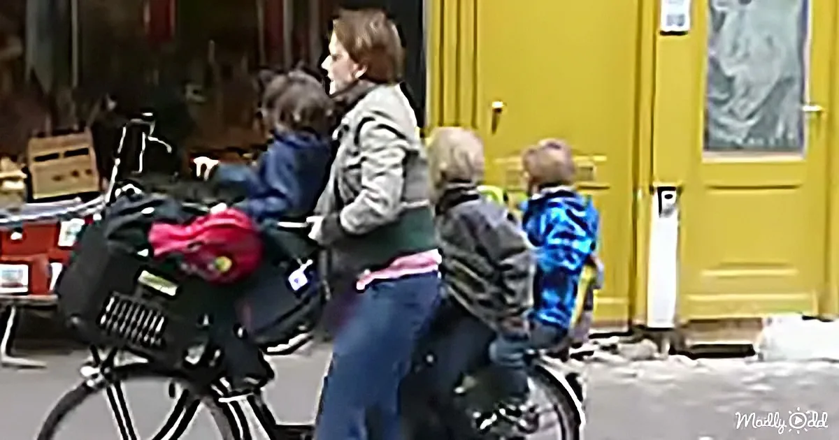 A woman walks with three children, two on bikes and one on a bicycle, near a yellow door.
