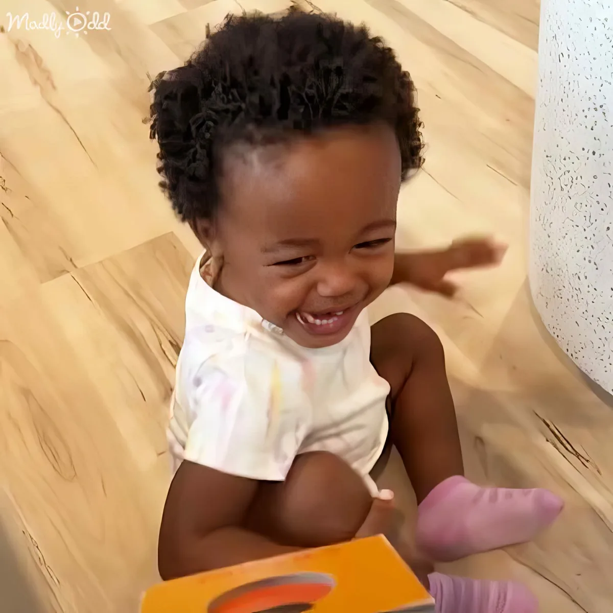 A baby with curly hair smiles while sitting on a wooden floor, holding a book.