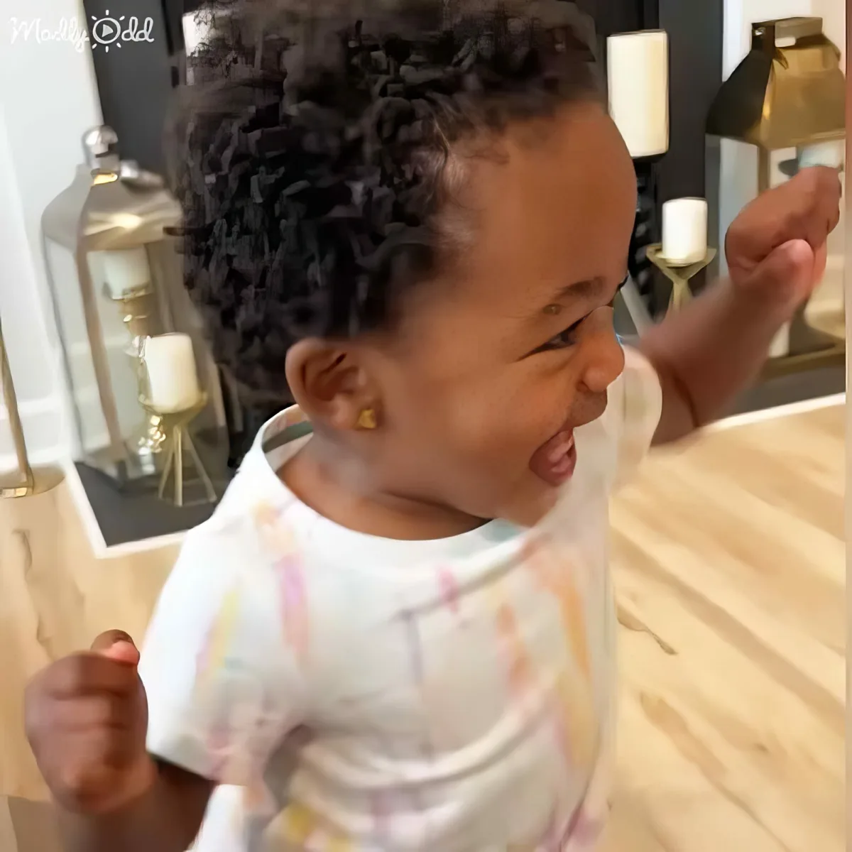 A baby with curly hair, wearing a white shirt with colorful patterns, is smiling and raising one hand.