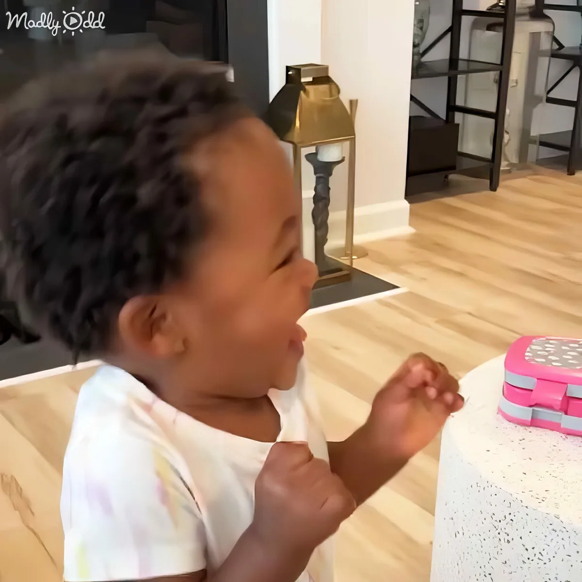 A baby in a high chair claps near a pink lunchbox on a table with a lace tablecloth.