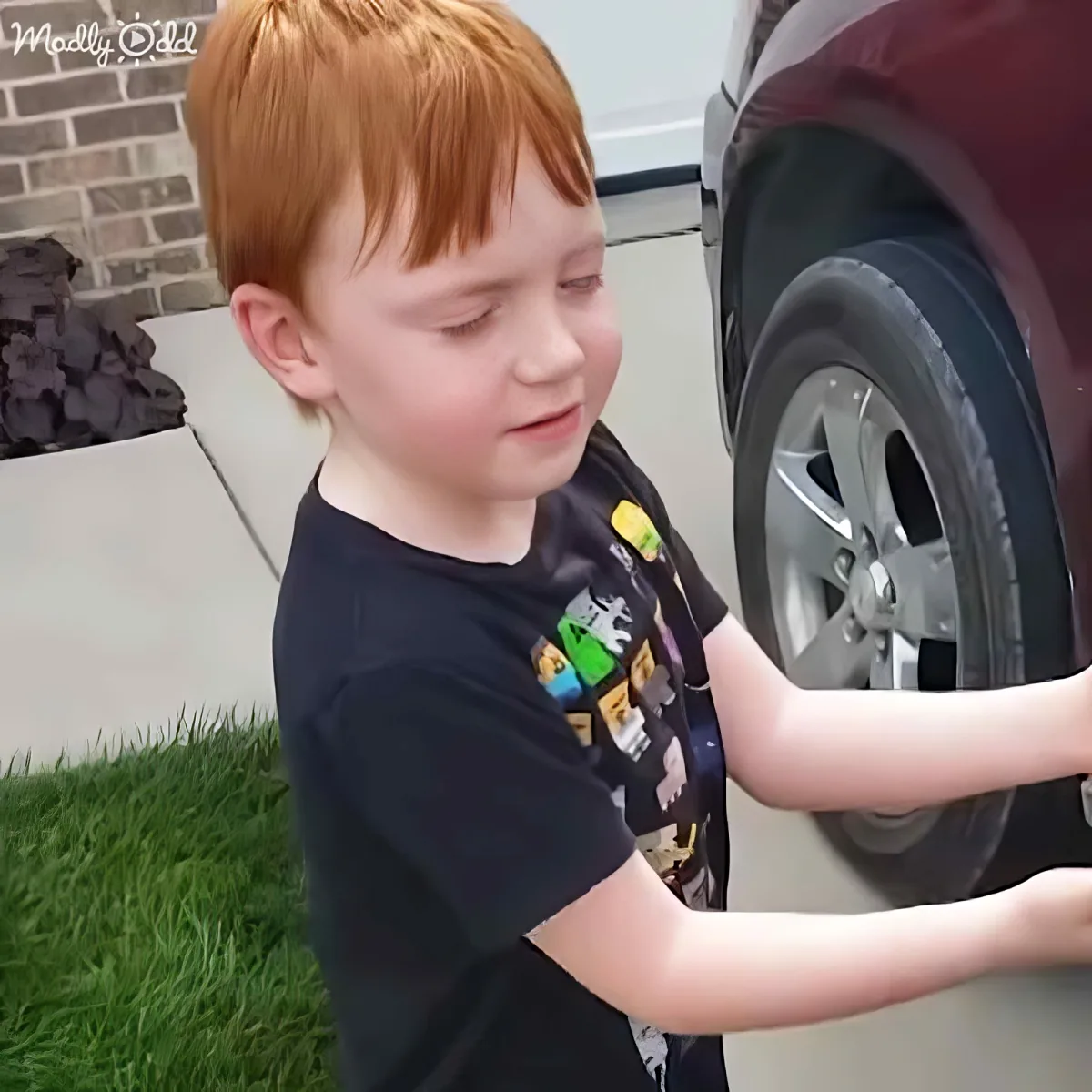 A young boy with red hair, wearing a black shirt, stands next to a car, looking down at the ground.