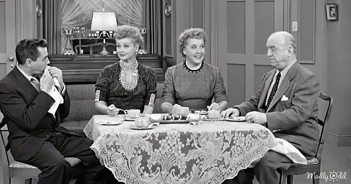 Four people sit around a table with tea sets, dressed formally, in a vintage setting.