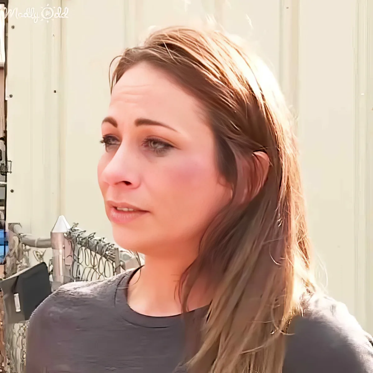 A woman with long brown hair, wearing a gray shirt, looks to the side with a neutral expression.