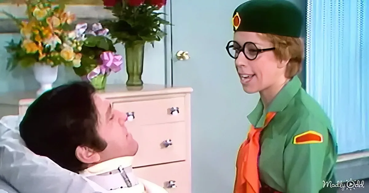 A person in a green uniform and glasses stands beside a person in a hospital bed, with a dresser and flowers in the...