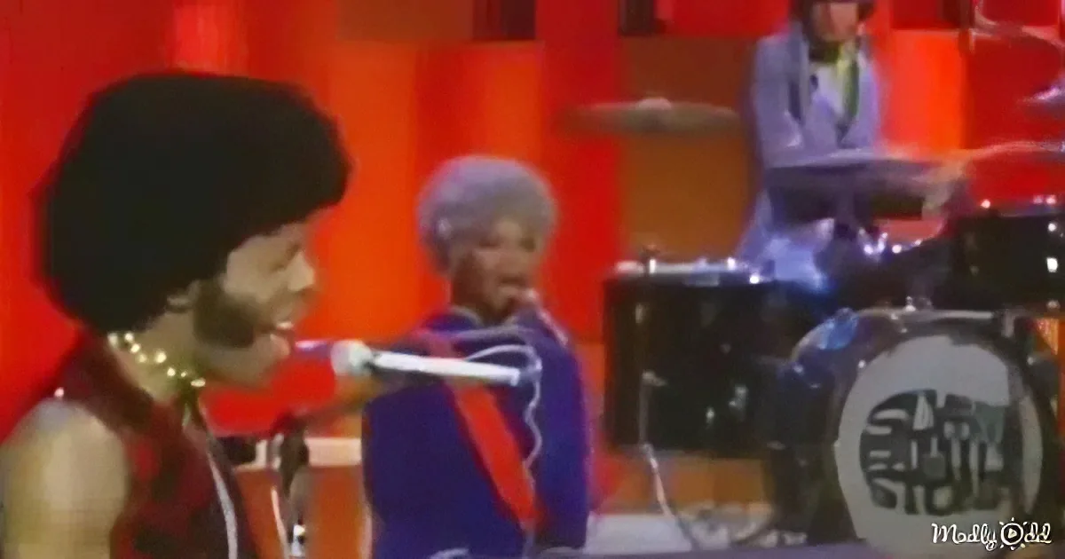A person in a red outfit is holding a microphone, with a drummer in the background on a stage.