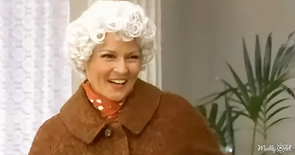 A smiling woman in a brown coat and white wig stands indoors near a plant.