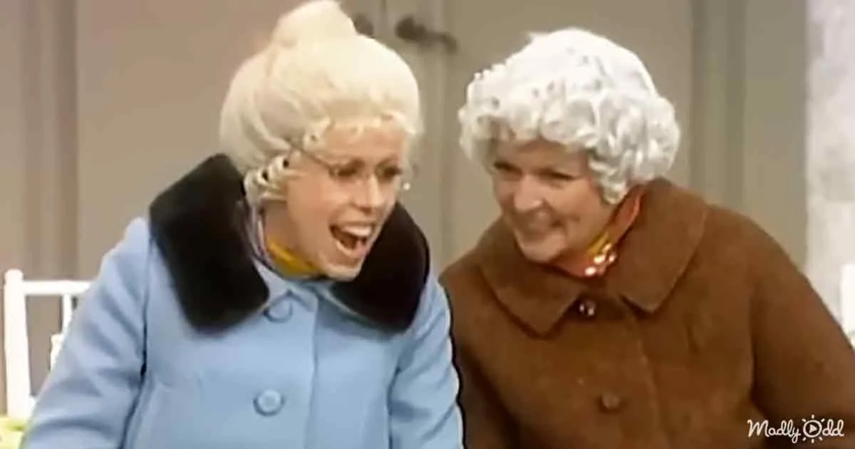 Two women in old-fashioned attire, one in blue, both with white wigs, appear to be laughing or reacting to something amusing.