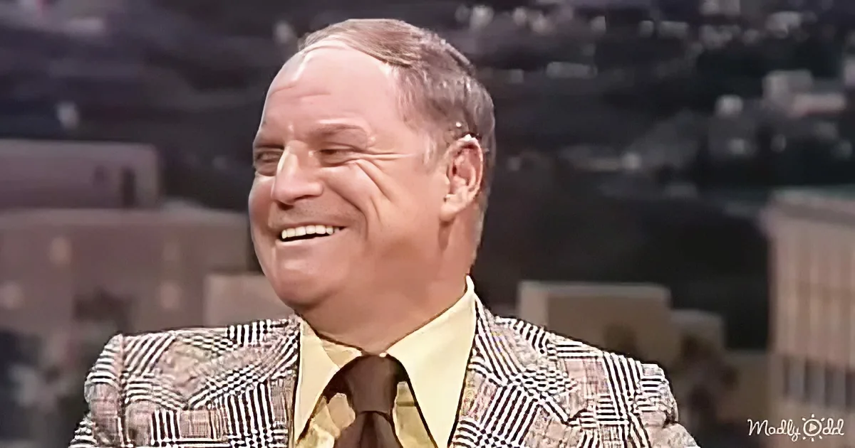 A man in a patterned suit and yellow shirt is smiling, with a blurred background suggesting a studio setting.