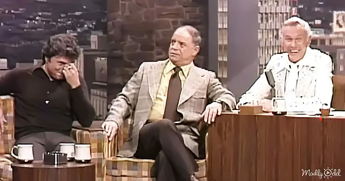 A man in a suit sits between two others, one laughing and the other smiling, in a talk show setting.