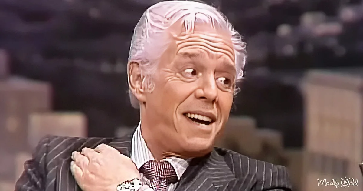 An older man in a suit and tie is smiling and gesturing with his hand.