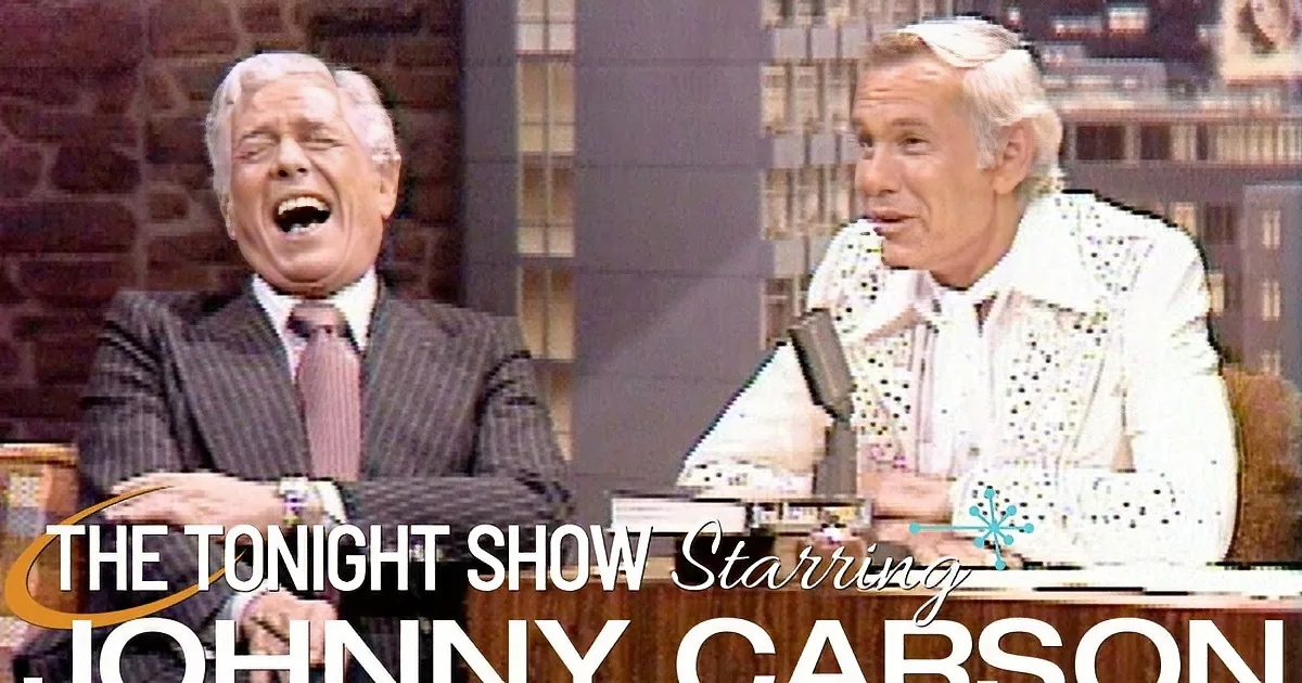 Two men, one in a suit and tie, the other in a white shirt, are seated and laughing on a talk show set.