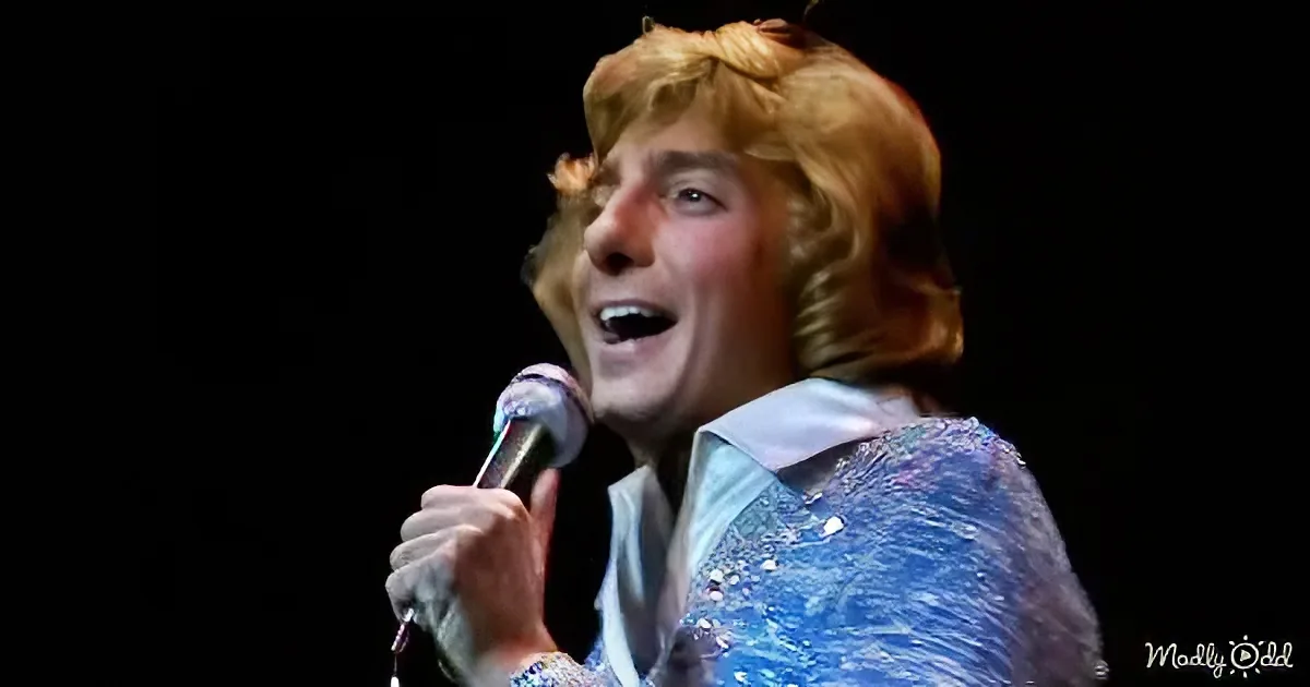 A person with blonde hair, wearing a blue sequined jacket, sings into a microphone against a dark background.