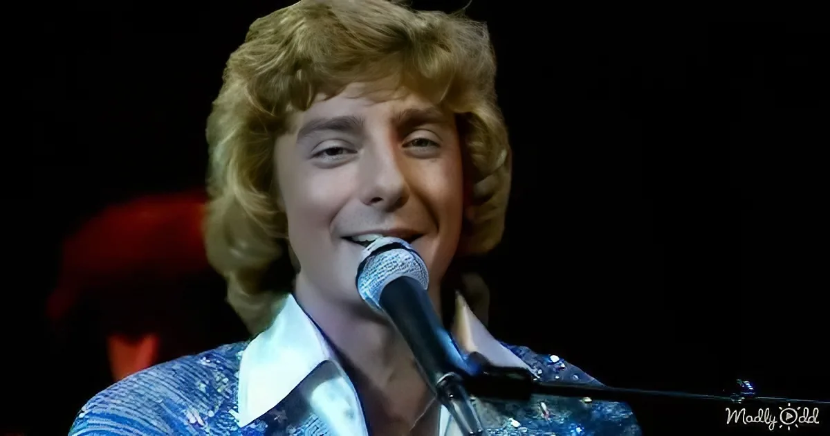 A person with curly blonde hair sings into a microphone, wearing a blue patterned shirt.