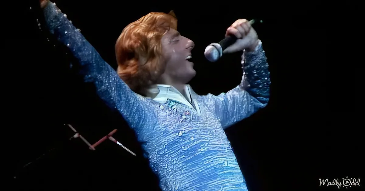 A person with red hair, wearing a sparkly blue outfit, sings passionately into a microphone with arms raised.