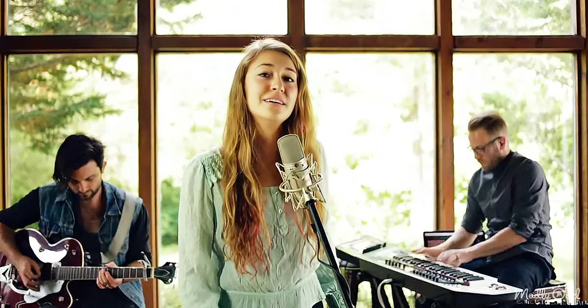 A woman sings into a microphone, accompanied by a guitarist and keyboardist in a bright, window-filled room.