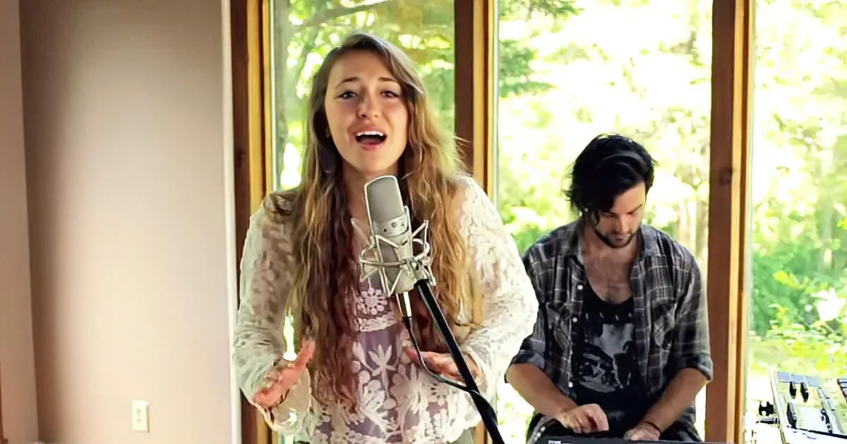 A woman sings into a microphone, wearing a floral top, with a man in a plaid shirt playing music in the background.