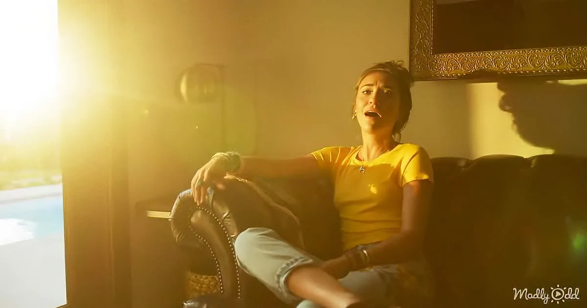 A woman in a yellow shirt sits on a couch, bathed in golden sunlight, with a pool and framed artwork in the background.