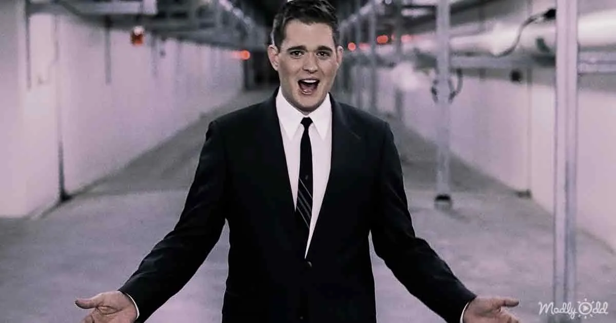A man in a black suit and tie stands in a dimly lit, industrial-looking hallway with his arms outstretched.
