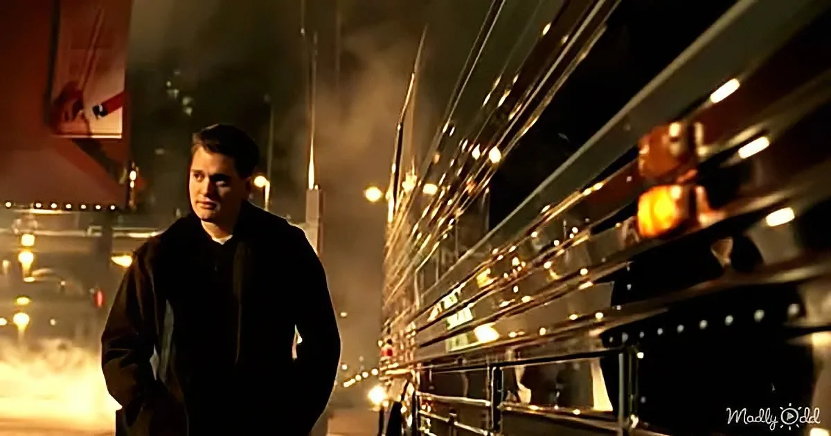 A man in a dark coat stands beside a large, illuminated bus at night.