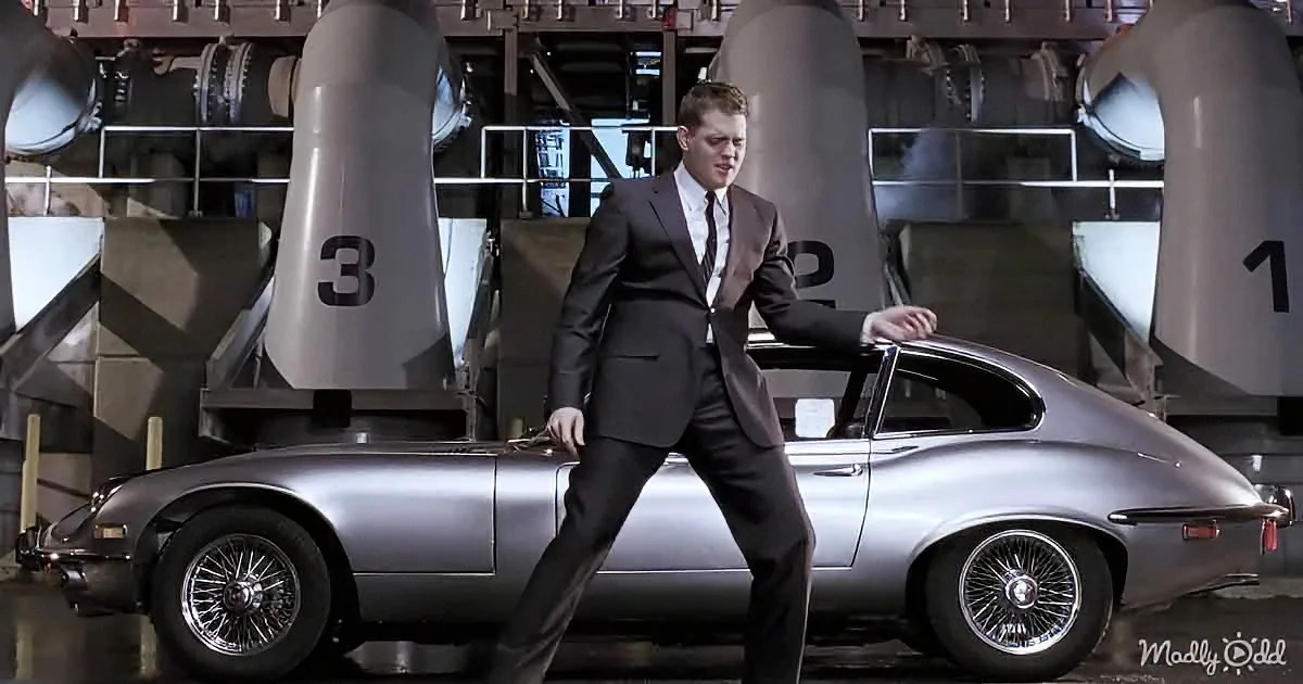 A man in a suit stands confidently beside a sleek silver sports car in a high-tech facility.