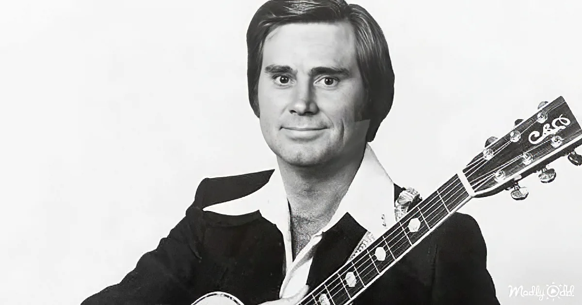 A man in a suit holds a guitar, smiling, against a plain background.