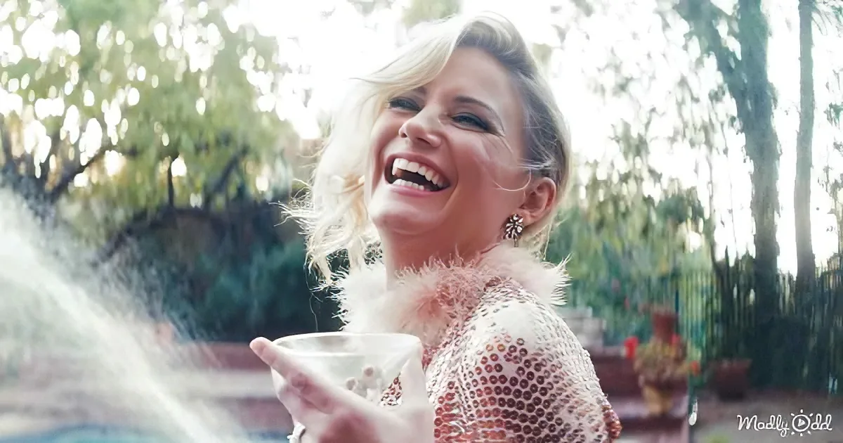 A woman with blonde hair, wearing a sequined dress with a fur collar, smiles while holding a glass, with a fountain and...