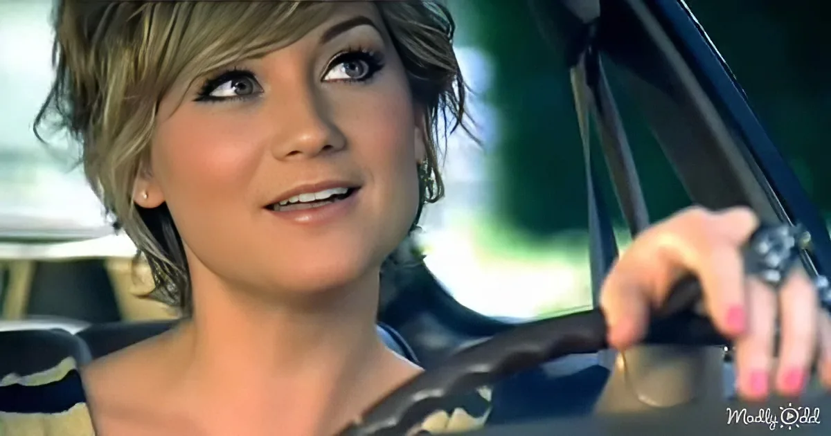 A woman with short blonde hair drives a car, her hand on the steering wheel, looking up and smiling.